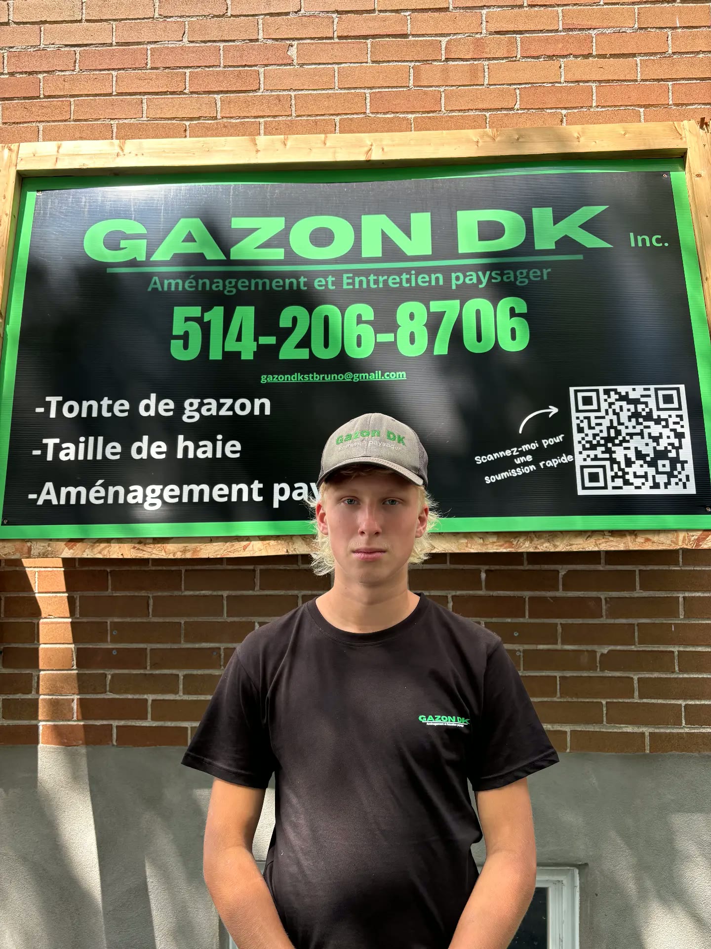 Denis, copropriétaire de Gazon DK, en t-shirt noir devant l'affiche de l'entreprise.