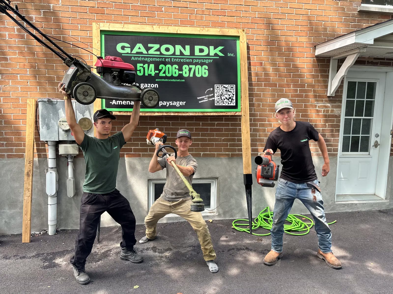 Les trois copropriétaires de Gazon DK posant avec une tondeuse, un coupe-bordure et un souffleur devant l'affiche de l'entreprise.